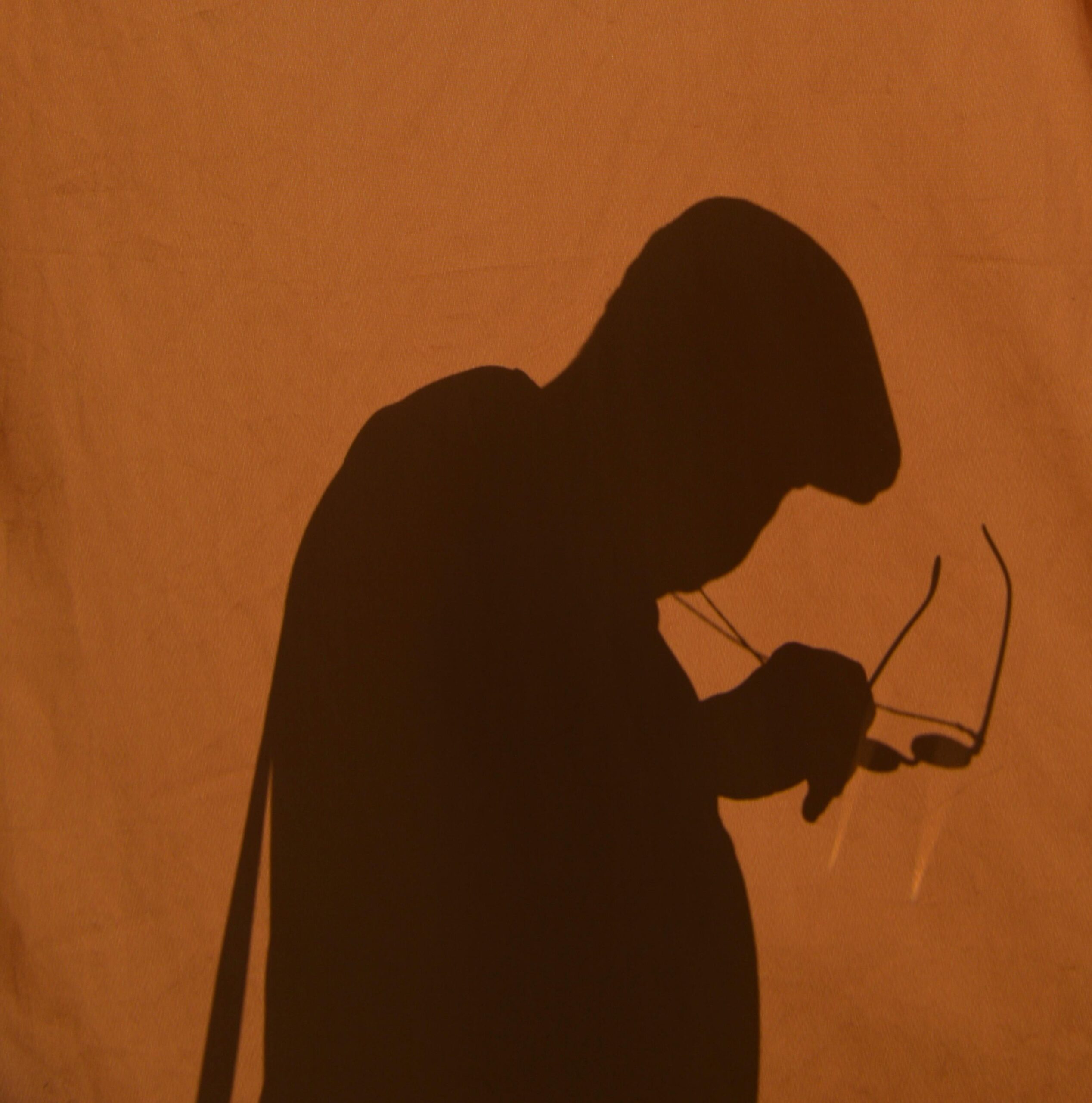 A shadowy silhouette of a person in a hat, holding glasses against a warm, sand-colored background.